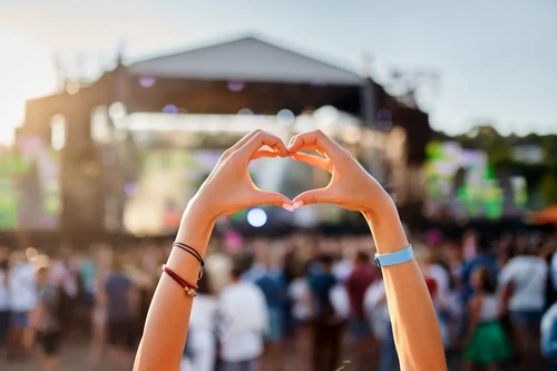 A concertgoer makes a heart shape with their hands at an outdoor music festival during the day.