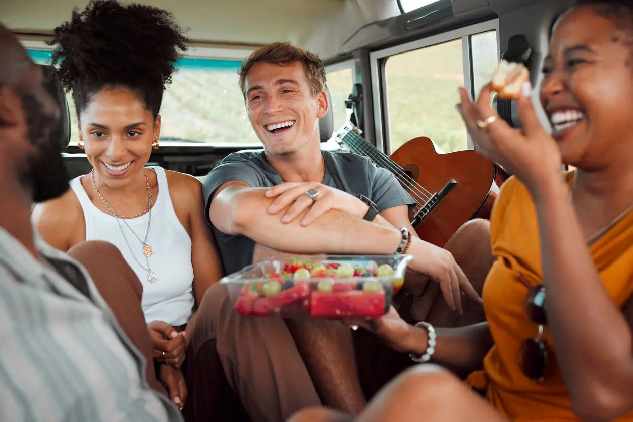 A group of friends sits in the back of a car, sharing fruit and laughing, with a guitar nearby during their road trip.