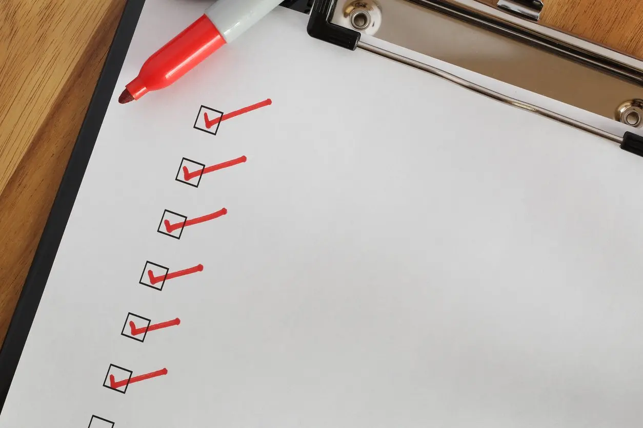 A red marker rests on a clipboard beside a checklist with several boxes marked with red checkmarks on a wooden desk. 